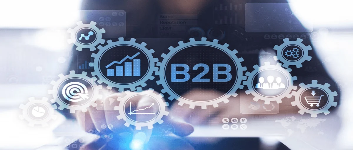 Tasarım- arka planda bilgisayar başında, klavyede bir tuşa basan bir çalışan (flu), ön planda çark tasarımları- birinin içinde "B2B" yazısı, diğerlerinin içinde iş grafikleri