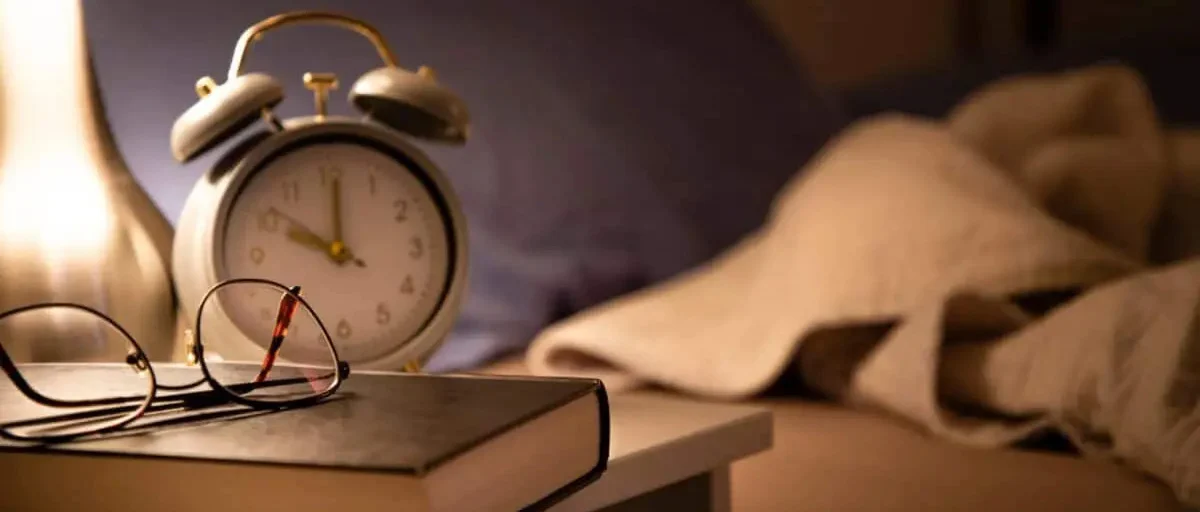 Başucunda duran kitap, üzerinde gözlük, yanında saat.